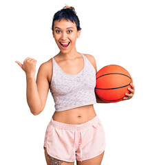 Sticker - Young woman holding basketball ball pointing thumb up to the side smiling happy with open mouth