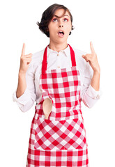 Poster - Beautiful young woman with short hair wearing professional cook apron amazed and surprised looking up and pointing with fingers and raised arms.