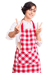 Poster - Beautiful young woman with short hair wearing professional cook apron smiling looking to the camera showing fingers doing victory sign. number two.