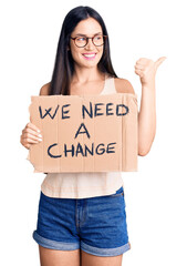 Poster - Young beautiful caucasian woman holding we need a change banner pointing thumb up to the side smiling happy with open mouth