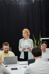 blonde businesswoman with digital tablet looking away near interracial colleagues working in office