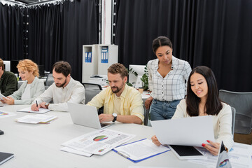 Wall Mural - smiling asian woman showing document to multiracial colleague while business team working at conference table