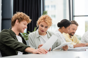 Wall Mural - smiling businesswoman showing document to colleague near interracial team on blurred background