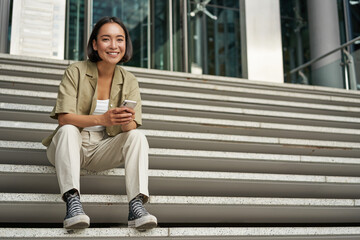 Wall Mural - Portrait of smiling asian girl sits on stairs outdoors, sending message, using smartphone app, looking at mobile phone screen