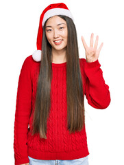 Wall Mural - Young chinese woman wearing christmas hat showing and pointing up with fingers number four while smiling confident and happy.