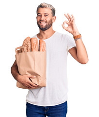 Poster - Young handsome blond man holding paper bag with bread doing ok sign with fingers, smiling friendly gesturing excellent symbol