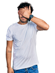 Wall Mural - Hispanic young man with beard wearing casual white t shirt covering eyes with hand, looking serious and sad. sightless, hiding and rejection concept