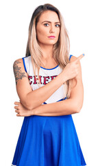 Canvas Print - Young beautiful blonde woman wearing cheerleader uniform pointing with hand finger to the side showing advertisement, serious and calm face