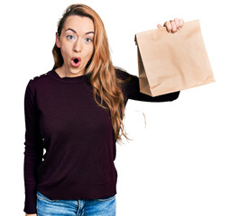 Canvas Print - Young caucasian woman holding take away paper bag scared and amazed with open mouth for surprise, disbelief face