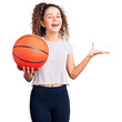 © Krakenimages.com - Beautiful kid girl with curly hair holding basketball ball celebrating victory with happy smile and winner expression with raised hands