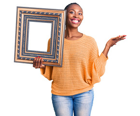 Wall Mural - Young african american woman holding empty frame celebrating victory with happy smile and winner expression with raised hands