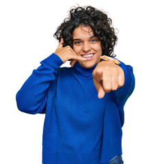 Canvas Print - Young hispanic woman with curly hair wearing turtleneck sweater smiling doing talking on the telephone gesture and pointing to you. call me.