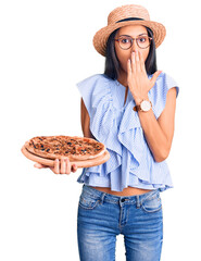 Sticker - Young beautiful latin girl wearing summer hat and glasses holding pizza covering mouth with hand, shocked and afraid for mistake. surprised expression