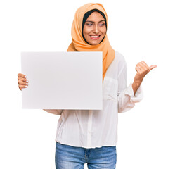 Poster - Young brunette arab woman wearing traditional islamic hijab holding banner pointing thumb up to the side smiling happy with open mouth