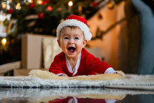 Happy Laughing Baby In Christmas Outfit Or Santa Claus Dress Crawling Infront Of Xmas Tree On A Blanket On The Floor In A Cozy Living Room 