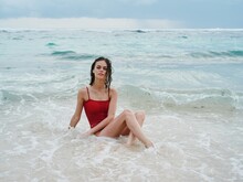 Woman Tourist In Red Swimsuit Sitting On The Sand On The Beach In The Ocean In The Waves, Cloudy Weather