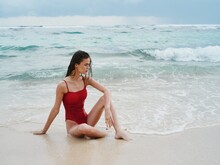 Woman Tourist In Red Swimsuit Sitting On The Sand On The Beach In The Ocean, Cloudy Weather