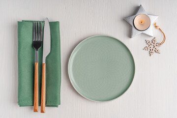 Top view of tableware set for one person served for christmas lunch or dinner celebration with green round plate, silver fork and knife, textile napkin and burning candle on white wooden background