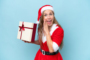 Wall Mural - Young blonde woman dressed as Mama claus holding a present isolated on blue background whispering something