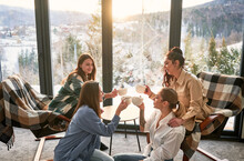 Young Women Enjoying Winter Weekends Inside Contemporary Barn House. Four Girls Having Fun And Clinking Cups Of Hot Tea Near Panoramic Windows At Sunset.