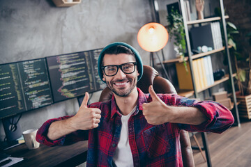 Photo of happy positive freelancer wear hat glasses showing two thumbs up smiling indoors workplace workstation loft