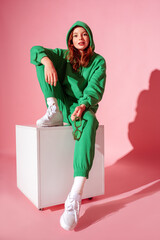 Wall Mural - Happy smiling fashionable freckled redhead girl wearing trendy green suit with hoodie, joggers,  white sneakers, sitting, posing on pink background. Full-length studio portrait
