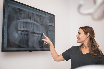 medicine, dentistry and health care concept - female dentist pointing to tooth showing x-ray of teeth on tv to patient at dental clinic office.