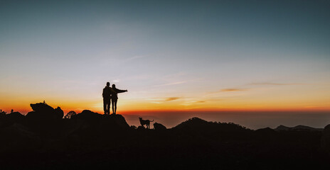 Wall Mural - Happy couple with dog embracing while standing on the top of the hill looking at sunset - Activities, love, travel, sport, nature and vacation concept