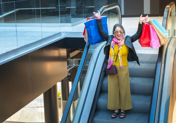 Senior lady celebrating with shopping bags on mechanic stairs at mall. Woman riding down escalator. High quality photo