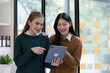 © NanSan - Cheerful asian business woman wearing sweater with colleagues working together at workplace female office worker during working day in office, looking tablet