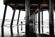 Monochromatic Pier Underside With A Surfer Walking Into The Sea. Abstract Geometry Of Huntington Beach Pier At Low Tide In California