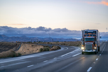 Brown classic big rig semi truck tractor with turned on headlight transporting cargo in reefer semi trailer driving on the straight one way road with fork intersection at twilight