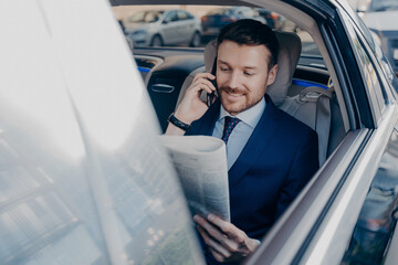 Good looking young executive manager in formal wear reads newspaper on backseat of car