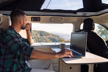 young man working on his laptop from his camper van in the middle of the nature, concept of freedom 