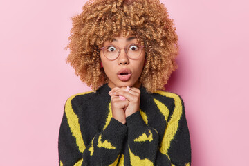 Wall Mural - Portrait of stunned woman with curly blonde hair keeps mouth opened gasps from wonder keeps hands under chin wears big transparent eyeglasses and casual jumper isolated over pink background.