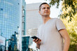 © Sergio - Smiling handsome caucasian young male holding mobile phone while standing on city street and looking away