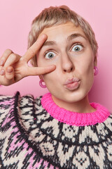 Poster - Vertical shot of surprised short haired woman keeps lips rounded makes peace gesture over eye looks wondered at camera wears winter jumper isolated over pink background. Body language concept