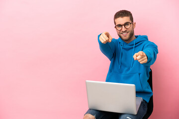 Poster - Young man sitting on a chair with laptop points finger at you while smiling