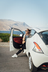 A beautiful brunette in sunglasses sits by a white car against the backdrop of the Mediterranean Sea and mountains