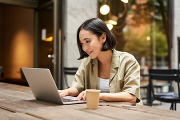 Wall Mural - Young asian woman, digital nomad working remotely from a cafe, drinking coffee and using laptop, smiling