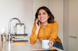 © Prostock-studio - Smiling young arab woman talking on cellphone and drinking coffee in kitchen