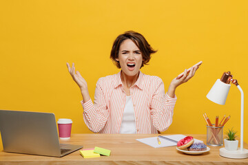 Wall Mural - Young indignant annoyed sad employee business woman wear casual shirt sit work at office desk hold mobile cell phone spread hands isolated on plain yellow color background. Achievement career concept.