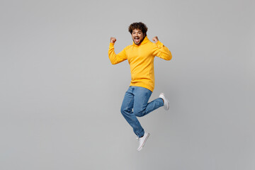 Full body successful lucky fun young Indian man 20s he wearing casual yellow hoody jump high do winner gesture clench fist isolated on plain grey background studio portrait. People lifestyle portrait.