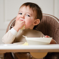 Wall Mural - A funny child is eating a grated apple with his mouth full while sitting on a kitchen chair. Hungry baby boy shoves food in his mouth, humor. Kid aged one year and three months