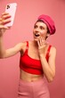 © SHOTPRIME STUDIO - A young woman blogger with colored pink hair and a short haircut takes a picture of herself on the phone and broadcasts a smile in stylish clothes and a hat on a pink background monochrome style