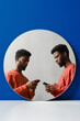 © Clique Images - Vertical creative studio mirror reflection double portrait of handsome young Black man wearing coral sweater using Internet on smartphone