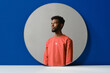© Clique Images - Horizontal contemporary creative studio shot of stylish young Black man wearing coral sweater looking away, round mirror reflection, blue background