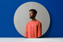 Horizontal Contemporary Creative Studio Shot Of Stylish Young Black Man Wearing Coral Sweater Looking Away, Round Mirror Reflection, Blue Background