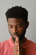 © Clique Images - Vertical close-up studio portrait of modern young African American man with beard on face wearing pink T-shirt posing on camera with praying hands gesture, gray background