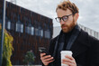 © YURII MASLAK - A young red-haired guy, a businessman in glasses, against the backdrop of urban style, checks mail on a smartphone, communicates in a messenger and drinks coffee. Online communication.